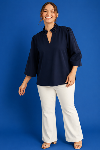A woman modeling a navy blue blouse with three-quarter sleeves and a split neckline, paired with white wide-leg pants, against a solid blue studio background.