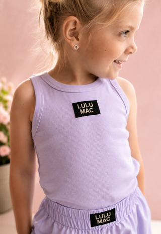 A young girl stands outdoors on a sunlit walkway wearing a light lavender sleeveless tank top and matching shorts
