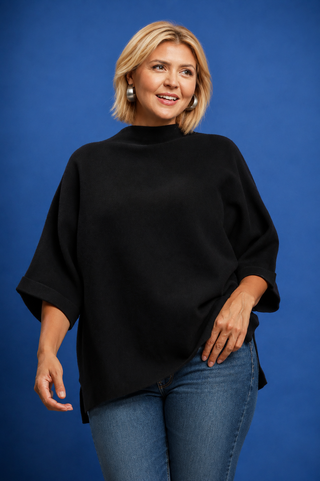 A blonde woman wearing a black oversized mock-neck top and light blue denim shorts, posing against a solid blue studio background.