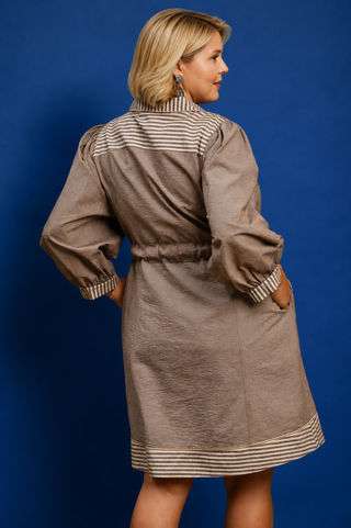 A woman modeling a brown linen-like dress with beige and brown striped details on the collar, cuffs, and hem, shown from a three-quarter back view against a solid blue background.
