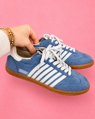 Blue sneakers with white stripes held by a hand against a pink background