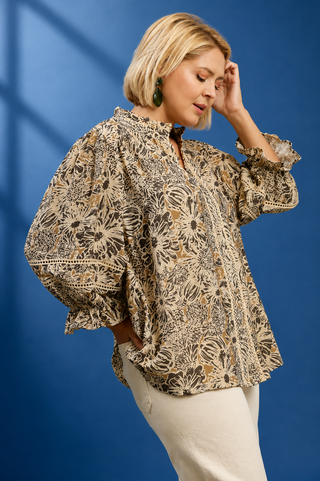 A woman modeling a loose, long-sleeve blouse with a beige, brown, and black floral block print, paired with cream pants, set against a solid blue studio background.