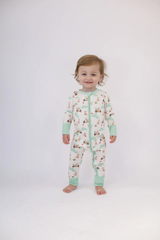 A toddler stands barefoot against a plain light gray studio background wearing a one-piece zip-up pajama with a white base, mint green cuffs and collar, and a scattered woodland animal print.