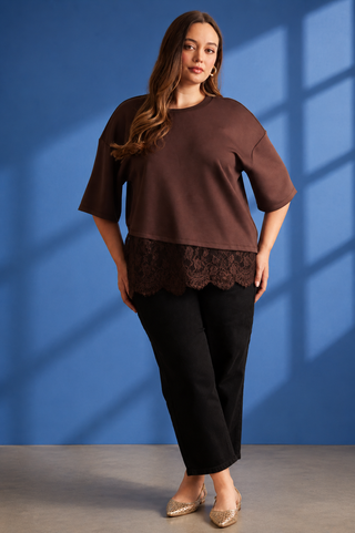 A woman models a brown short-sleeve top with a dark brown lace hem overlay, paired with black pants and beige pointed flats, set against a blue wall with window shadow patterns.