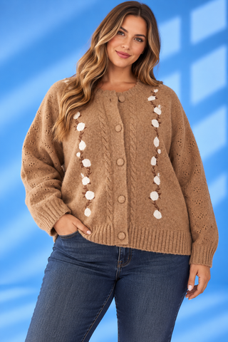 A woman wearing a brown cable-knit cardigan with white pom-pom accents down the front, paired with blue jeans