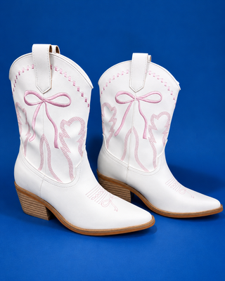 A pair of white leather cowboy boots with pink embroidered bows and hearts, featuring light brown stacked wood-look he
