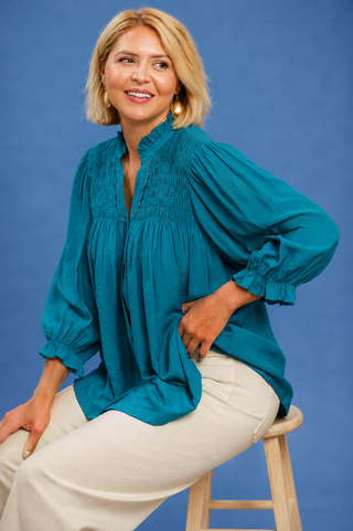 A woman with blonde hair sits on a wooden stool wearing a teal smocked blouse and cream pants against a solid blue studio background.