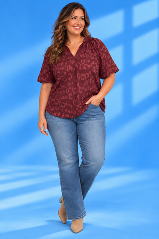 A woman modeling a short-sleeve burgundy top with a subtle leopard print pattern, paired with light-wash blue jeans and beige heels
