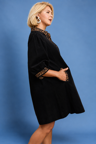 A woman modeling a black velvet-like short dress with brown and black patterned trim at the collar and sleeve cuffs, posed against a solid blue studio background.
