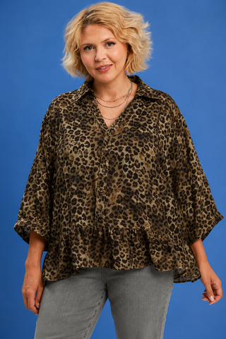 A woman wearing a leopard-print blouse with wide three-quarter sleeves and a peplum hem, paired with gray jeans, standing against a solid blue background.