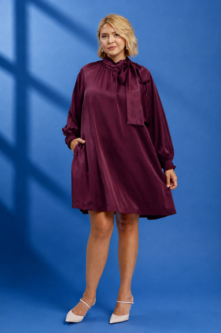 A woman modeling a satin-like burgundy long-sleeve dress with a high neck, bow detail, and side pockets, standing against a blue studio backdrop with window shadow overlays.
