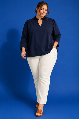A woman modeling a navy blue long-sleeve blouse with a split V-neckline, paired with white pants and brown slide sandals, set against a solid blue studio background.