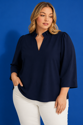 A woman modeling a navy blue blouse with a split V-neckline and elbow-length sleeves, paired with white pants, set against a solid blue studio background.