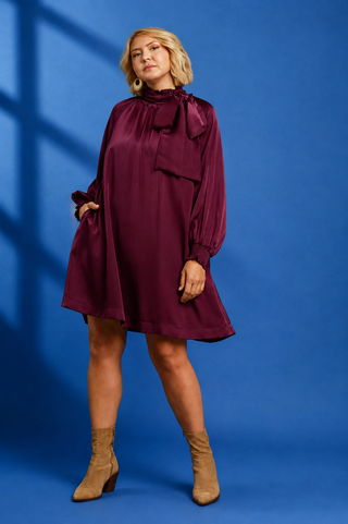 A woman modeling a long-sleeve satin-like mini dress in deep burgundy with a large bow at the high neckline, paired with tan ankle boots, against a blue studio backdrop.