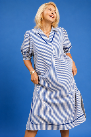 A woman modeling a blue and white vertically striped midi dress with dark blue piping details, posed against a solid blue studio background.