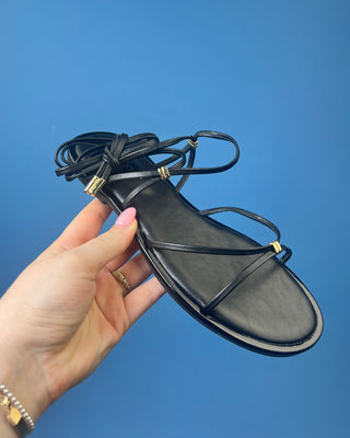 A pair of black leather flat sandals with thin strappy details and small gold hardware, against a solid blue background.