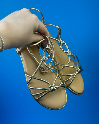 A pair of gold metallic strappy sandals with a tan footbed, against a solid blue background.