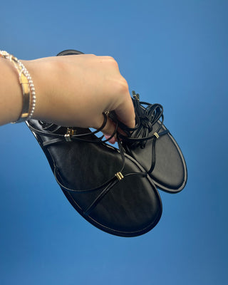 A pair of black leather flat sandals with thin strappy details and small gold hardware, against a solid blue background.