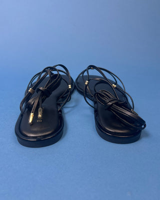 A pair of black leather flat sandals with thin strappy details and small gold hardware, against a solid blue background.