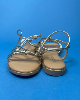 A pair of gold metallic strappy sandals with a tan footbed, against a solid blue background.