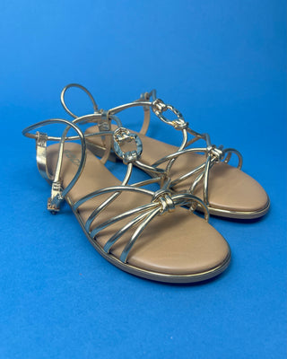 A pair of gold metallic strappy sandals with a tan footbed, against a solid blue background.