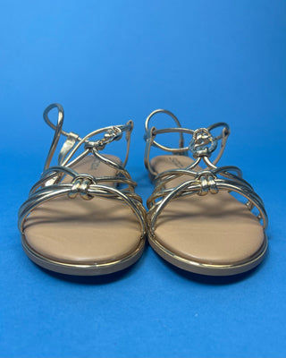 A pair of gold metallic strappy sandals with a tan footbed, against a solid blue background.