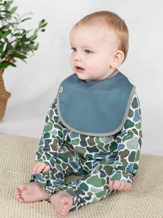A baby sitting on a textured beige mat wearing a long-sleeve footed onesie with a green and blue camouflage pattern and a slate blue bib.