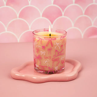 Candle in a glass jar with pink bow pattern on a pink heart-shaped tray against a pink background