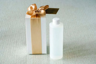 Clear bottle with white cap next to a gift box with a gold ribbon on a light gray background