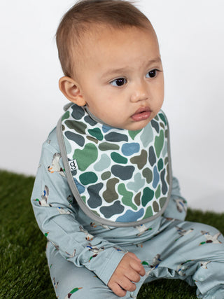 A baby wearing a light blue long-sleeve jumpsuit with a small bird print and a green, blue, and gray camouflage-patterned bi