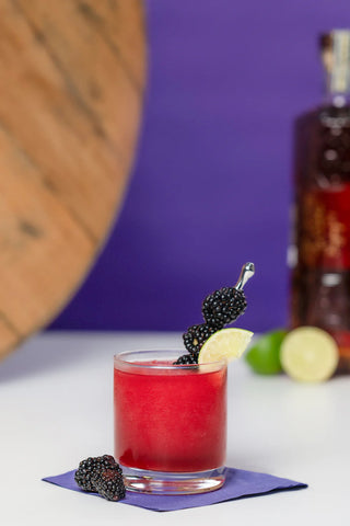 Red cocktail with blackberries and a lemon wedge on a purple napkin, with a bottle in the background.