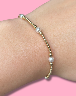 Gold and pearl bracelet on a wrist against a pink background
