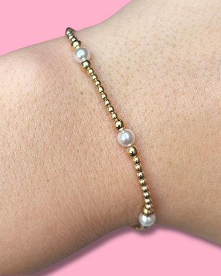Gold beaded bracelet with pearls on a wrist against a pink background