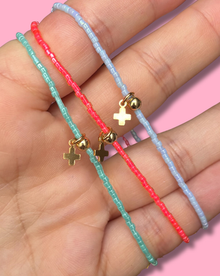 Three colorful beaded bracelets with gold cross charms on a hand against a pink background