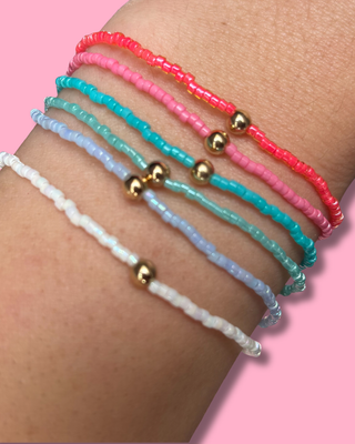Close-up of a wrist wearing three beaded bracelets with gold beads on a pink background