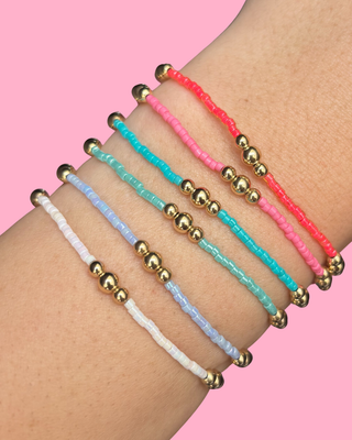 Three beaded bracelets in gold, pink, and blue on a wrist against a pink background