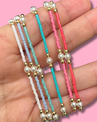 Four colorful beaded bracelets with pearl-like beads on a hand against a pink background