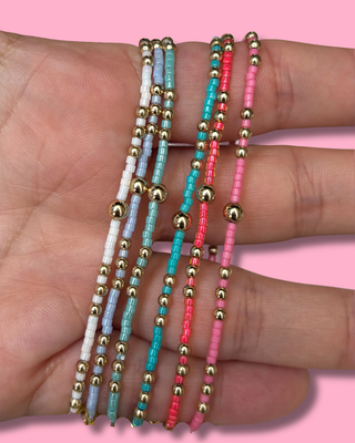 Three beaded bracelets in gold, blue, and pink on a hand with a pink background