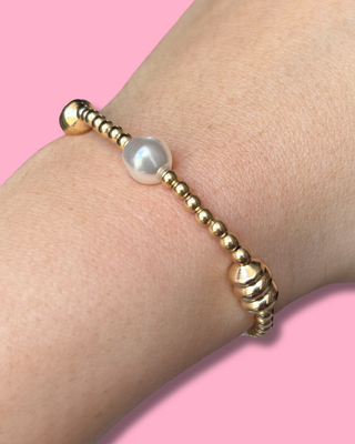Gold beaded bracelet with a pearl on a wrist against a pink background