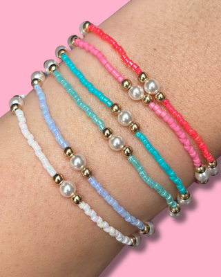 Three beaded bracelets in white, blue, and pink on a wrist against a pink background