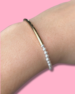 Gold bangle and pearl bracelet on a wrist against a pink background