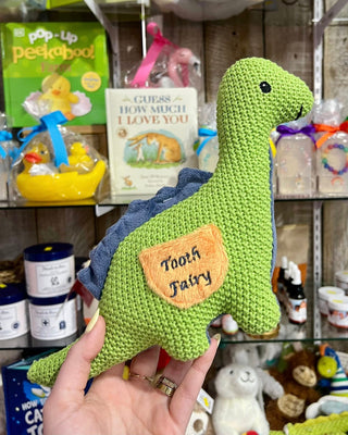 Green knitted dinosaur toy with 'Tooth Fairy' patch held in front of a store shelf.