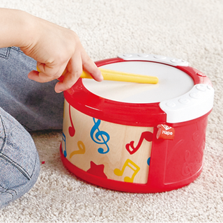 Child playing with a red toy drum featuring musical notes on a carpeted floor.