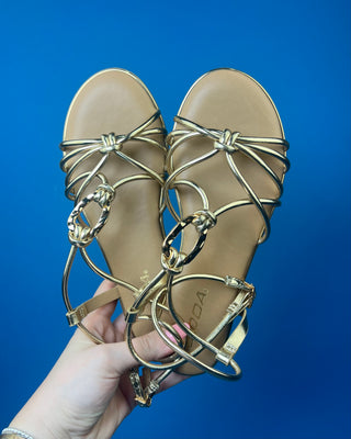 A pair of gold metallic strappy sandals with a tan footbed, against a solid blue background.