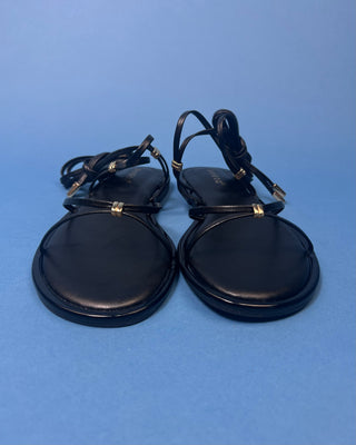 A pair of black leather flat sandals with thin strappy details and small gold hardware, against a solid blue background.