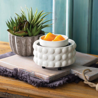 A white ceramic scented wax warmer with a textured bubble pattern and a small bowl of orange wax on top, styled on a wooden board with a potted succulent in the background.