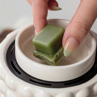 A close-up of a hand with gradient manicured nails holding a green square-shaped wax melt above a white ceramic scented candle warmer with a black inner ring.