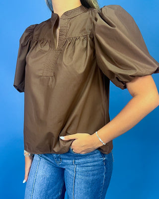 A woman wearing a brown short-sleeve blouse with puff sleeves and a split neckline, paired with blue jeans, set against a solid blue background.