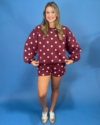 A woman modeling a matching burgundy and white polka dot sweatshirt and shorts set against a solid blue studio backdrop.