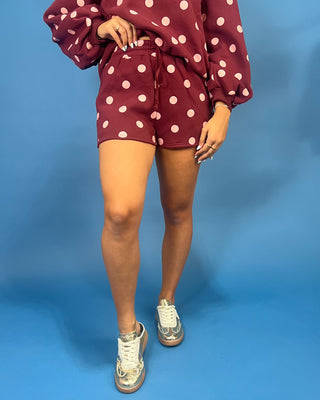 A person wearing a matching burgundy and white polka dot lounge set, featuring a long-sleeve top and shorts, paired with white sneakers, set against a solid blue background.